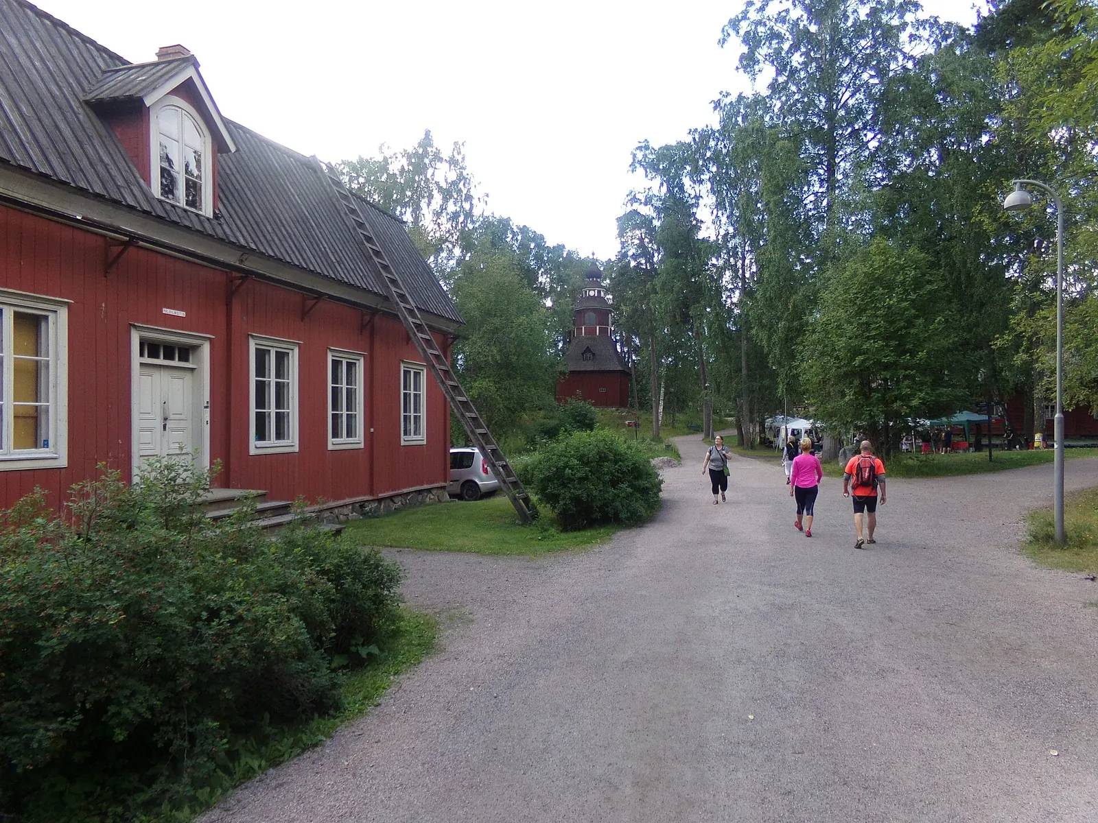 Hiekkatie ja punainen puutalo Seurasaaren ulkomuseossa, taustalla kellotapuli puiden keskellä