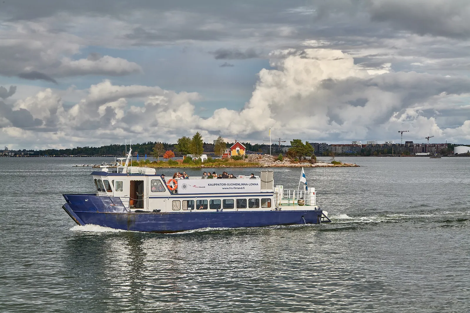 Kauppatori–Suomenlinna–Lonna-lautta kulkee Helsingin edustan merellä, taustalla pieni saari punaisine mökkeineen