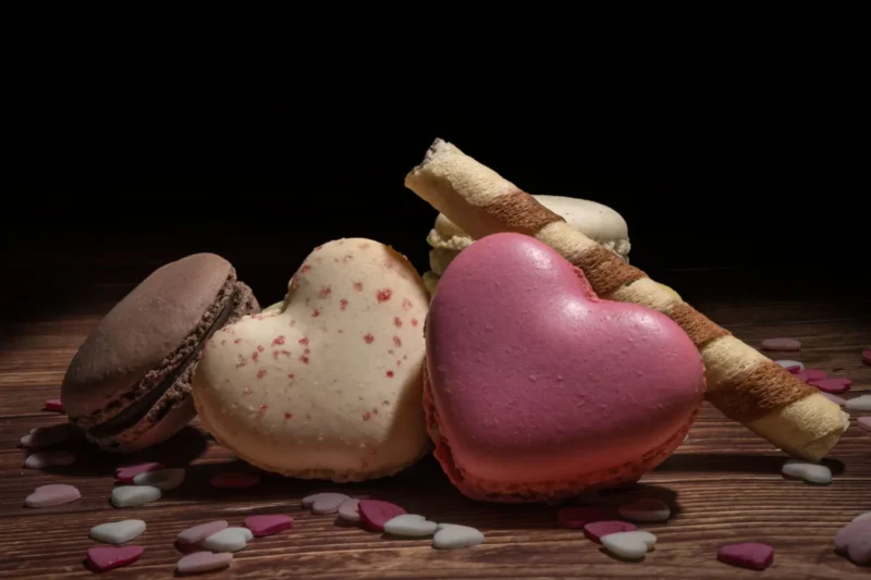 Sydämenmuotoisia vaaleanpunaisia ja vaaleita macaron-leivoksia puupöydällä sokerisydänten keskellä