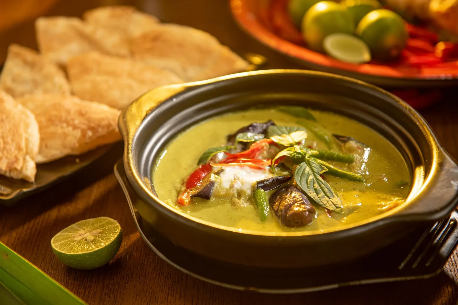 Thaimaalainen vihreä curry mustassa kulhossa, vieressä rotia-leipää ja lime