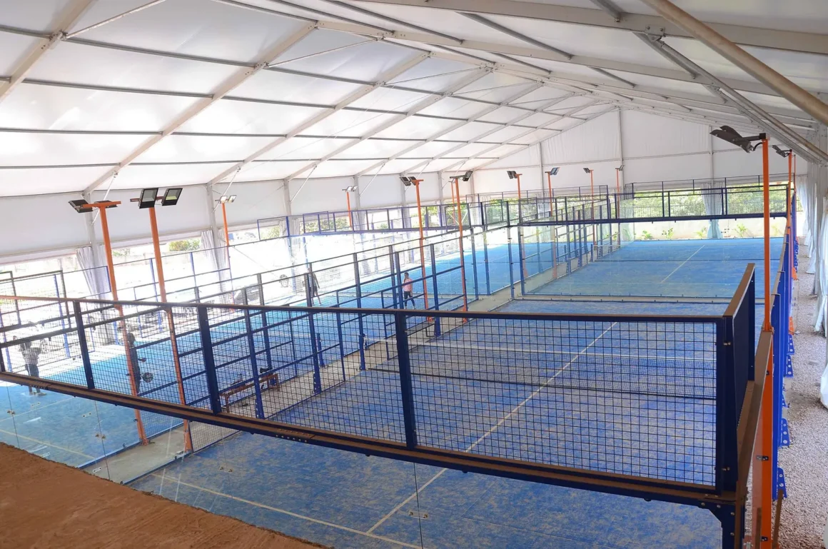 Useita sinisiä padel-kenttiä lasiseinineen valkoisen katoksen alla